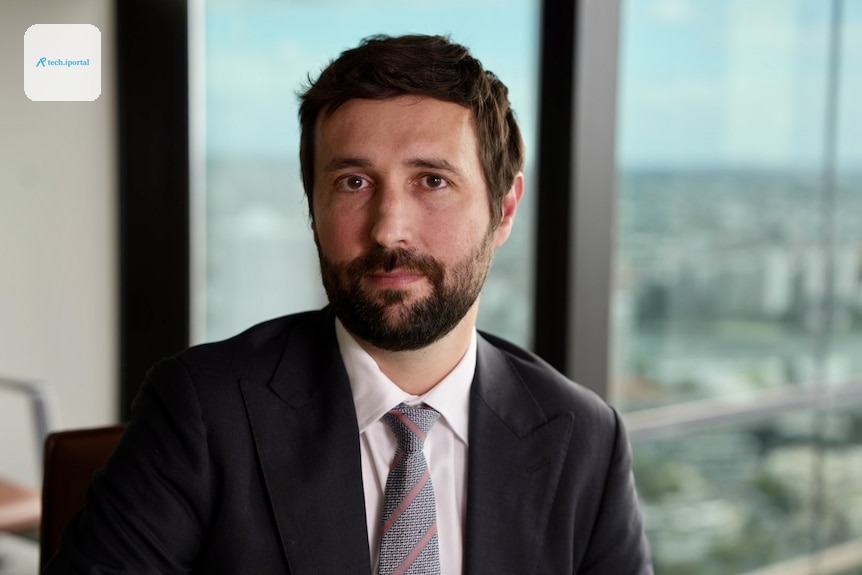 A man in a black suit and tie looks at camera.