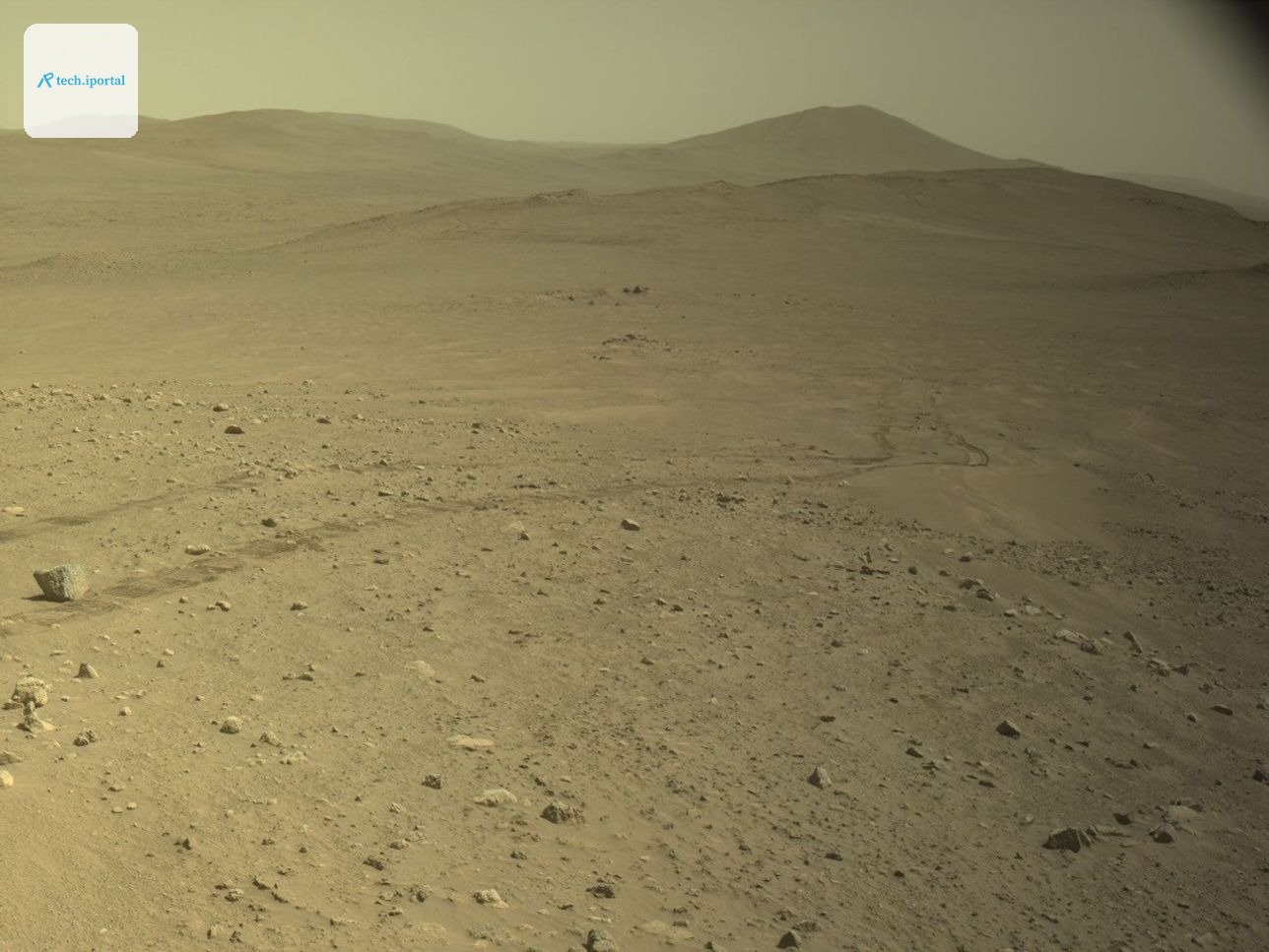 A wide view of the Martian surface taken by the Perseverance rover. The foreground features an arid, tan-colored terrain scattered with rocks and pebbles of varying sizes. Faint, parallel rover tracks