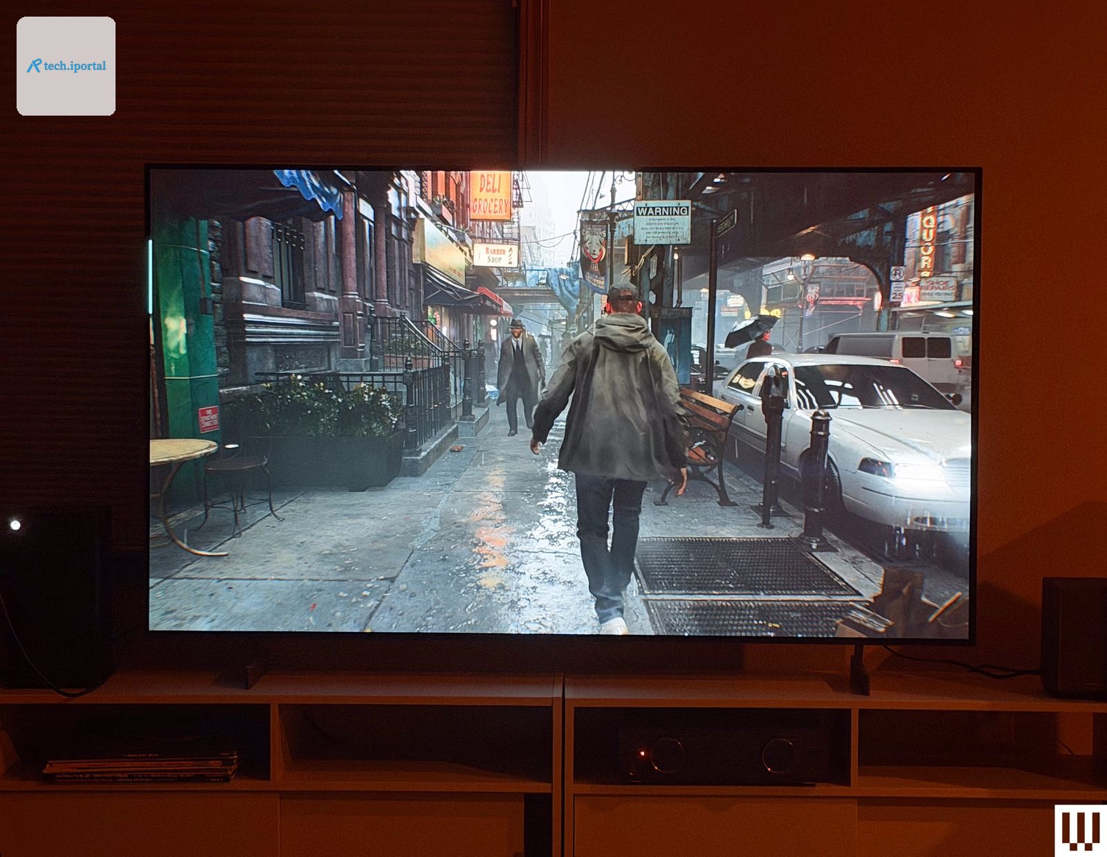 Flatscreen tv in a dark room with the screen showing a rainy city street with parked cars subway tracks overhead and...