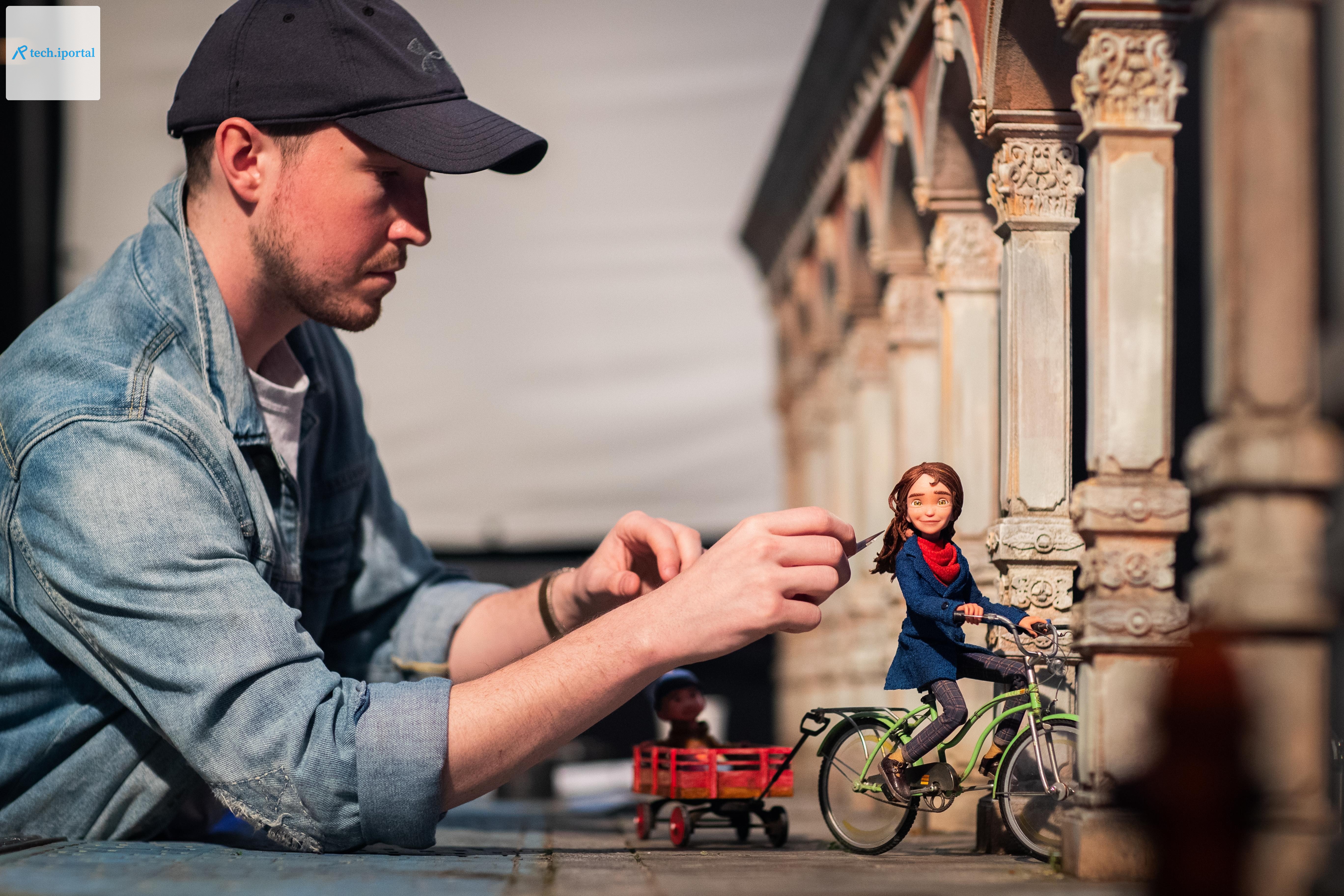 An animator uses a small tool on a puppet on a set for the stop-motion film Wildwood. The puppet is riding a bike near a building.