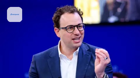 Reuters Dario Amodei hld the closed fingers of his left hand together as he mades a point. He is wearing a white shirt and blue jacket, black-rimmed glasses and has slightly curly hair. He is sat agai