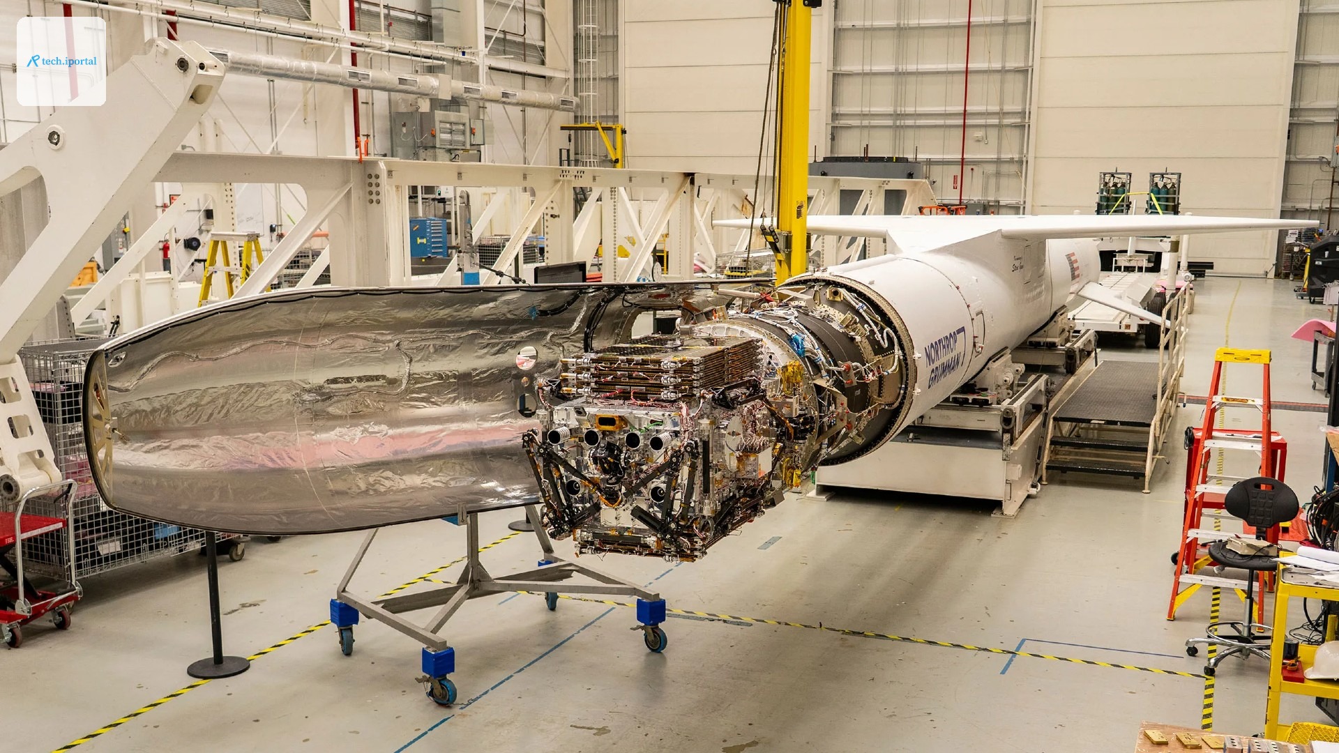 a long white rocket sits horizontal inside a hanger with its nose cone open, exposing a satellite with many components and actuators folded up into a dense rectangular shape