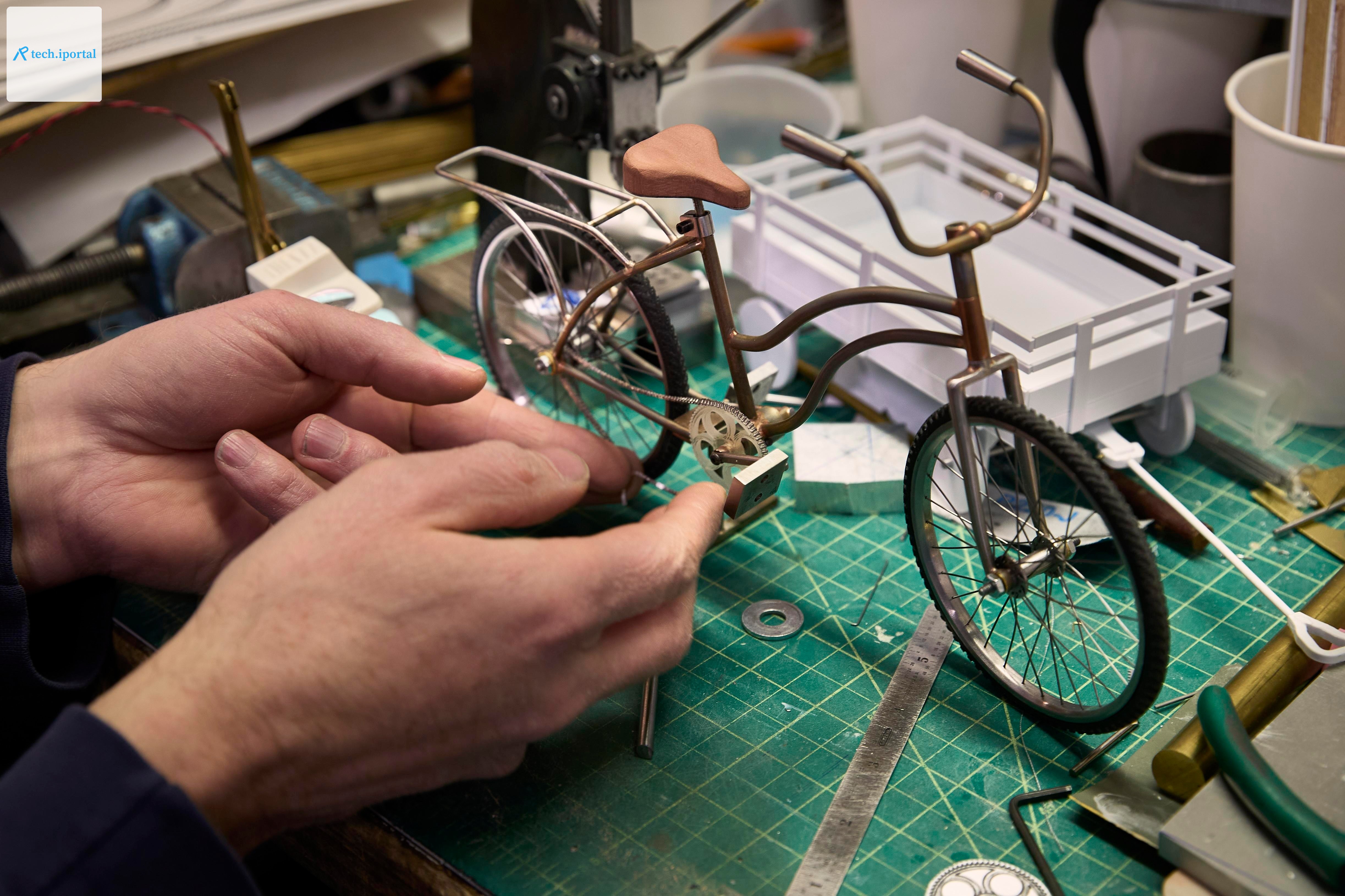 A pair of hands working on a model of a bicycle on a crafting table, with various tools and parts nearby.