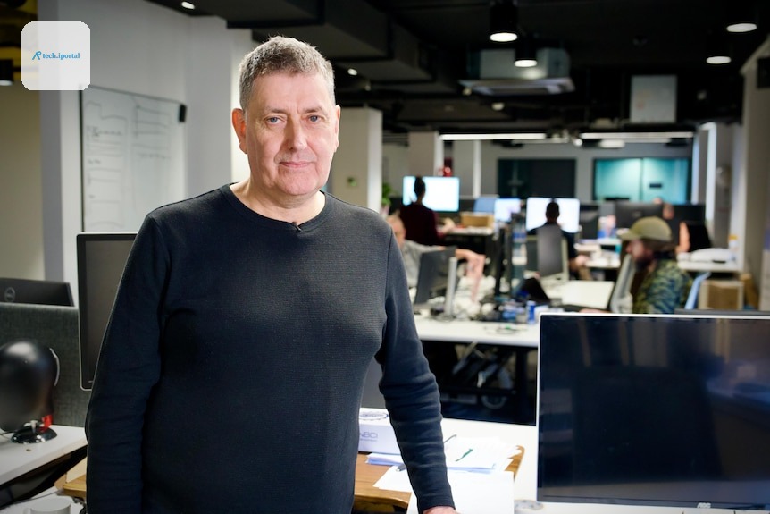 A man in a long black shirt stands in an open plan office with people and computers in the background.