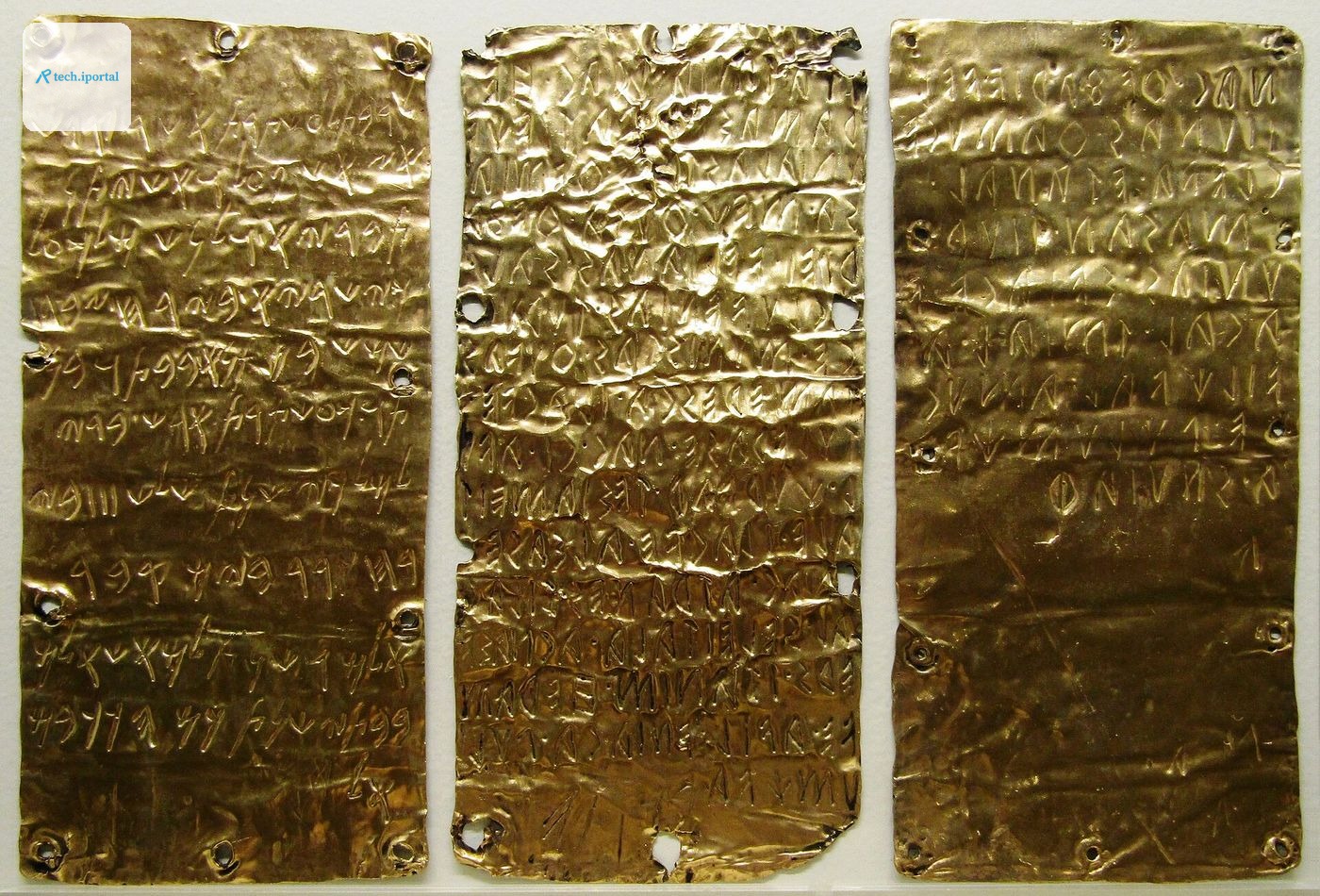 Three thin rectangular sheets of gold displayed side by side, each crumpled slightly and covered in rows of incised ancient lettering, with small nail holes around their edges.