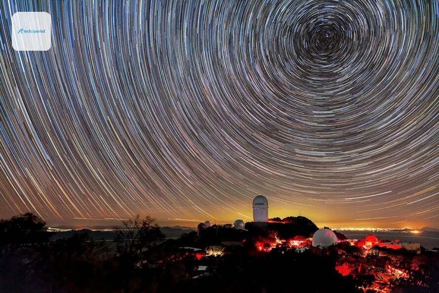 A long-exposure image captures circular star trails above telescope domes illuminated in red light on a dark mountain. A golden glow on the horizon marks distant city lights.