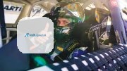 Racing driver gives a thumbs up from inside a car, wearing a helmet and safety gear.