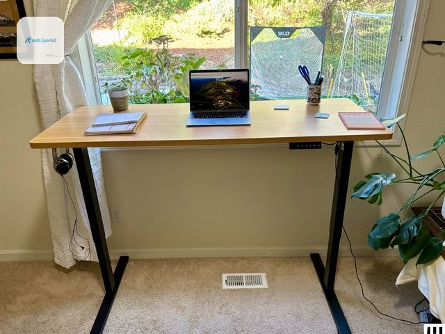 Flexispot EN1 Standing Desk a thin desk with light brown top and 2 black legs. On top is a spiral notebook small potted...